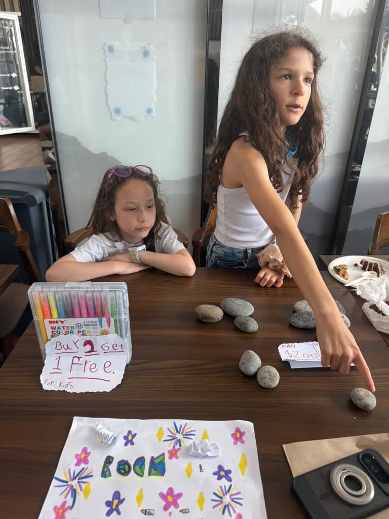 Two girls running a hand-painted rock stand with welcome and pricing signs