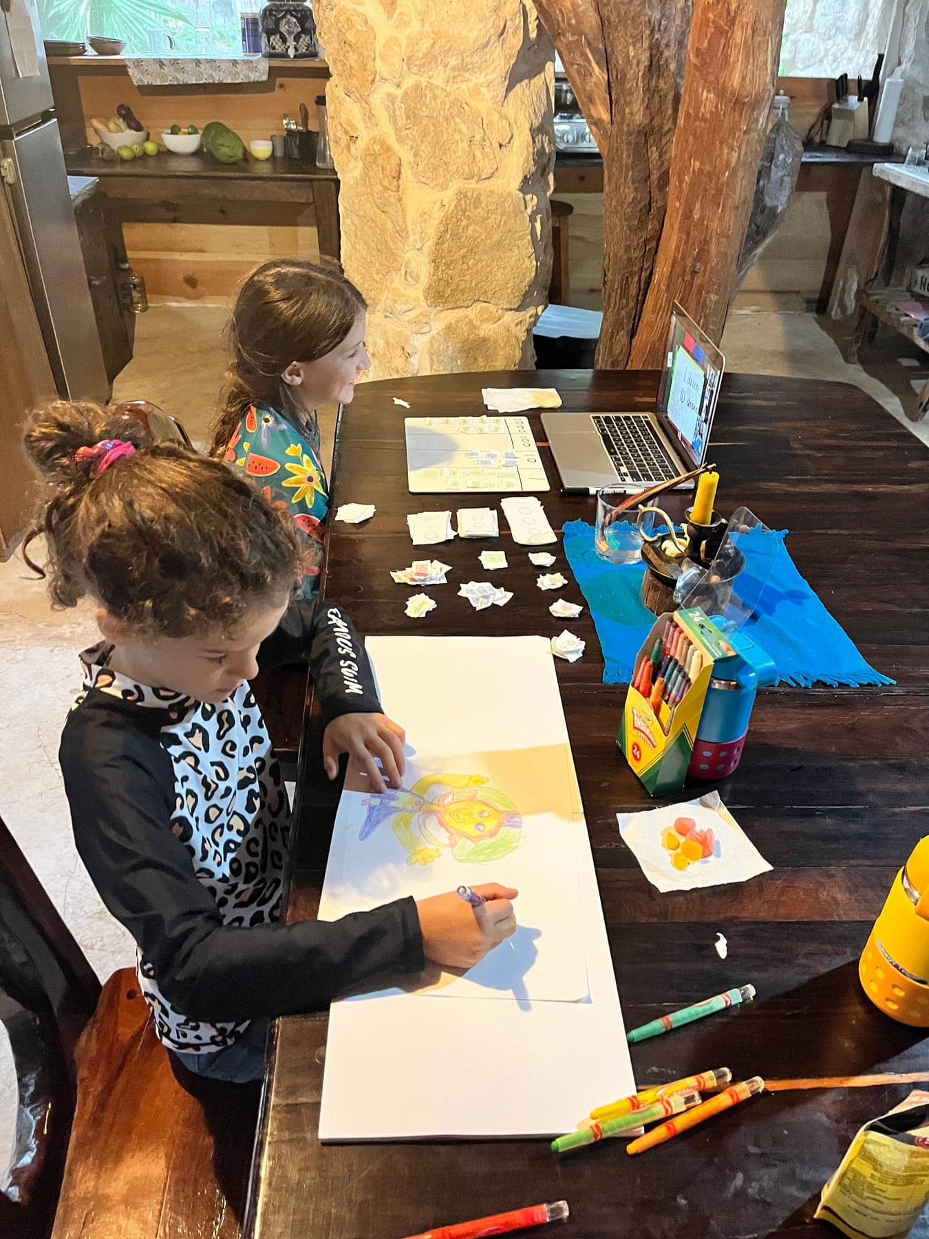 Two children drawing and working with paper money materials at a wooden table