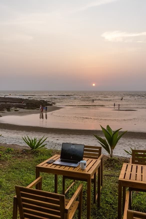 A laptop open on a beach table at sunset in Goa, the ocean glowing behind it